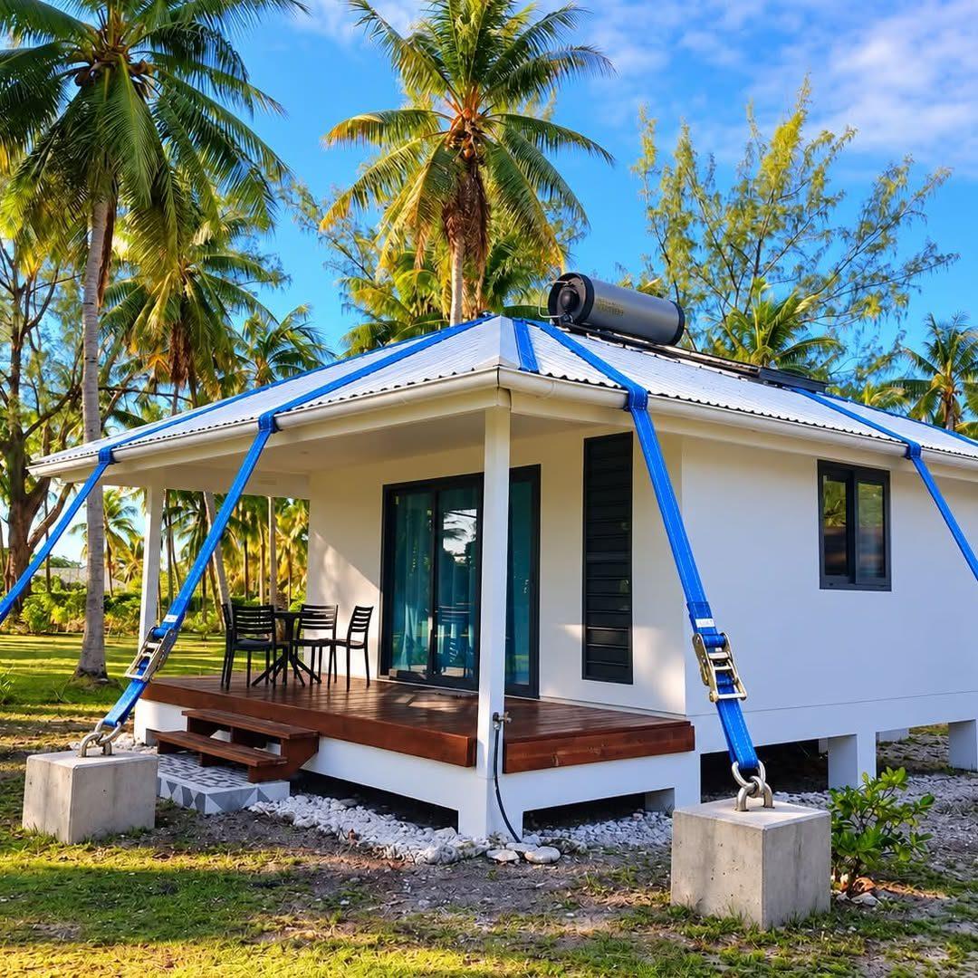 Maison en bois sous les cocotiers, toiture ancrée au sol par des sangles bleues tendues jusqu'à des blocs de béton